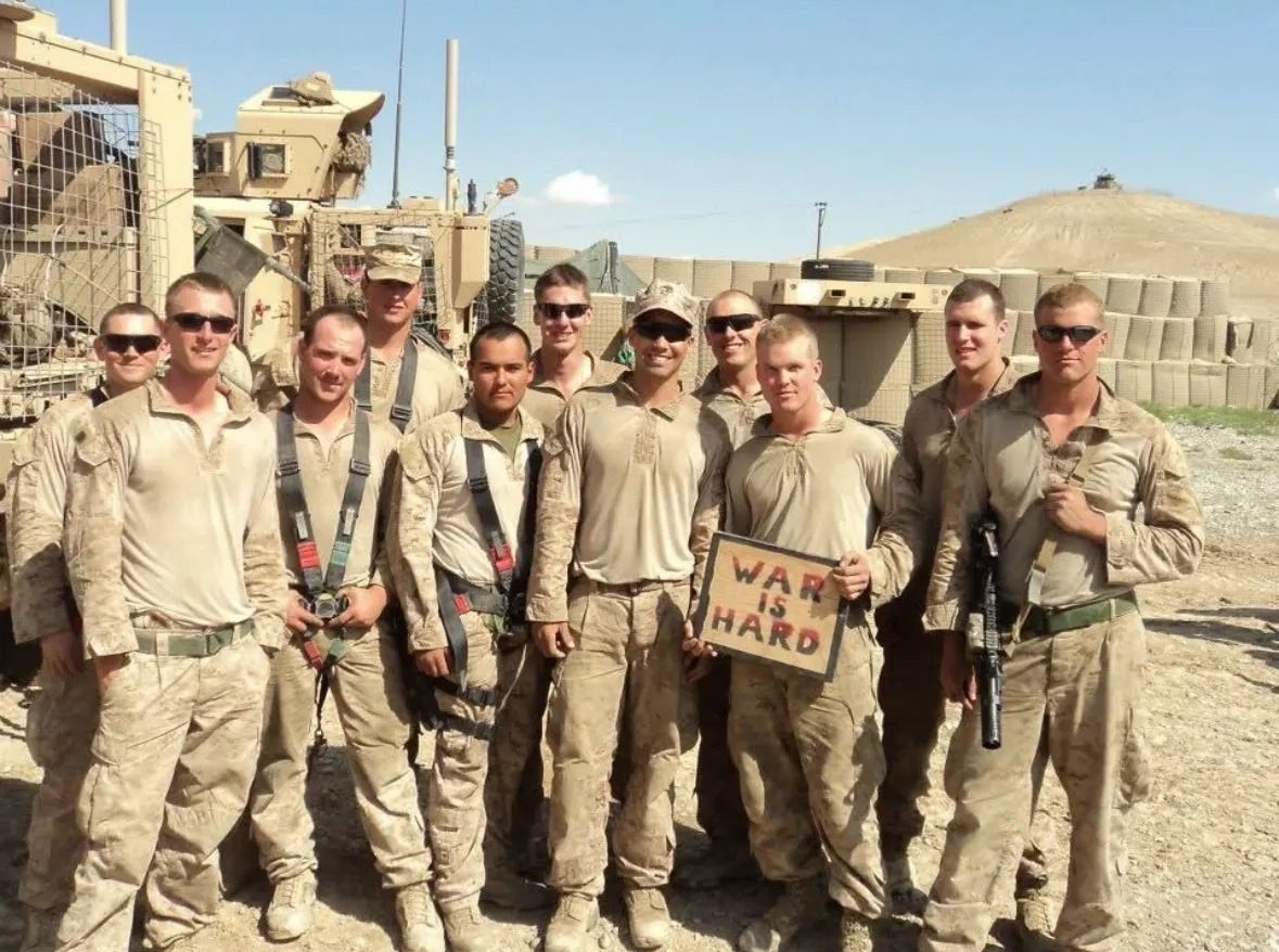 A group of Marines in desert utilities pose in front of an armored vehicle at a forward base; one holds a hand-painted sign reading “War is hard”