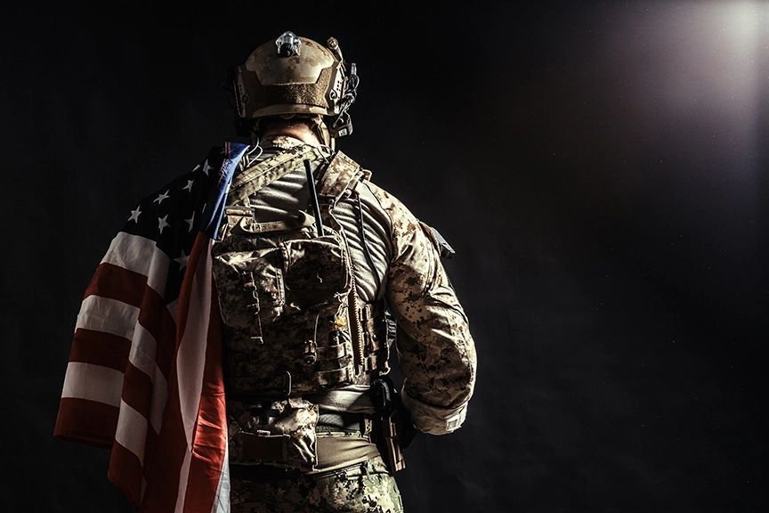 A soldier in combat uniform, seen from behind, with an American flag draped over one shoulder