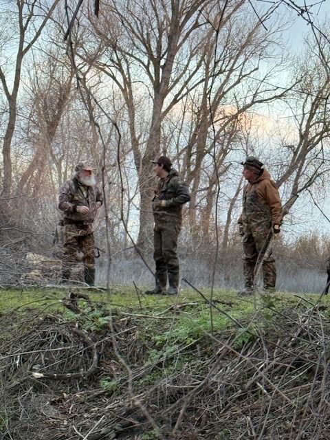 Three hunters in waders talk at the edge of a marsh at dawn beneath bare cottonwoods