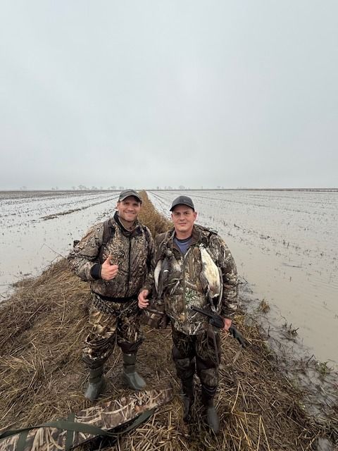 Two hunters in camouflage smile and hold up their harvested ducks in a rice field